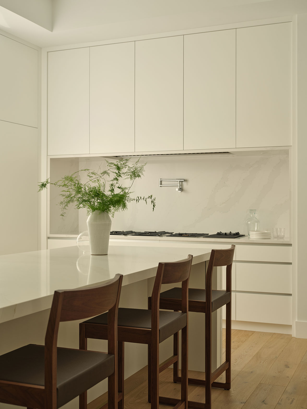 Modern kitchen with white cabinets and countertops, wooden bar stools, a potted plant on the island, and a minimalist backsplash with a faucet above the stove.