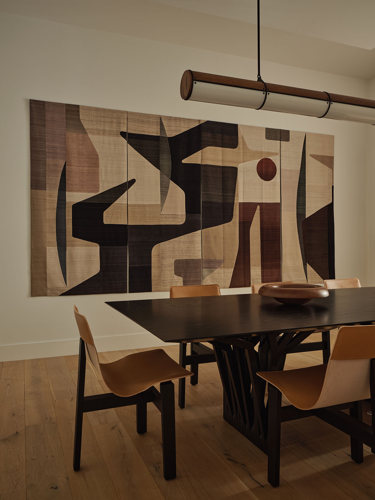 A modern dining room with a dark wood table, brown leather chairs, wooden flooring, and an abstract geometric wall tapestry in neutral and dark tones.