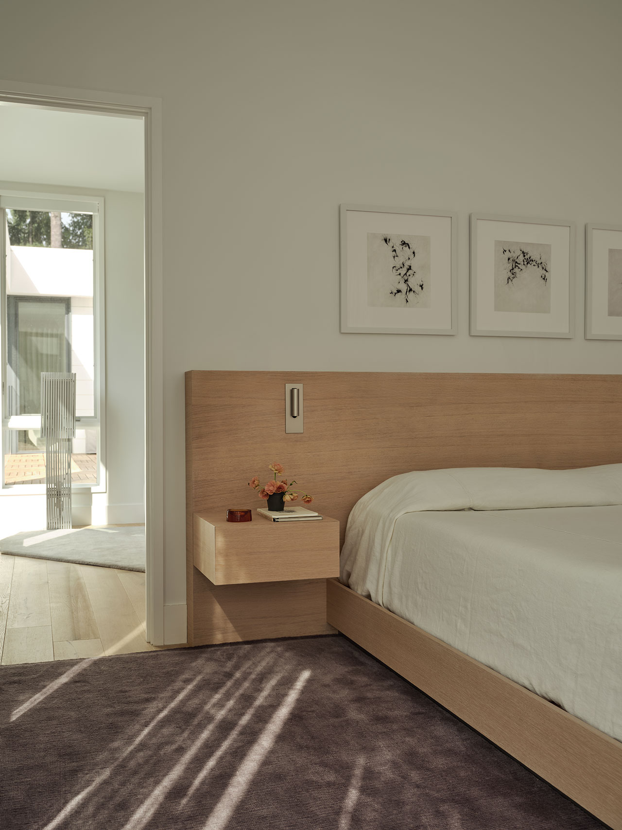 Minimalist bedroom with a light wood headboard, floating nightstand, white bedding, abstract wall art, and sunlight streaming across a purple carpet. Doorway opens to a bright adjacent room.