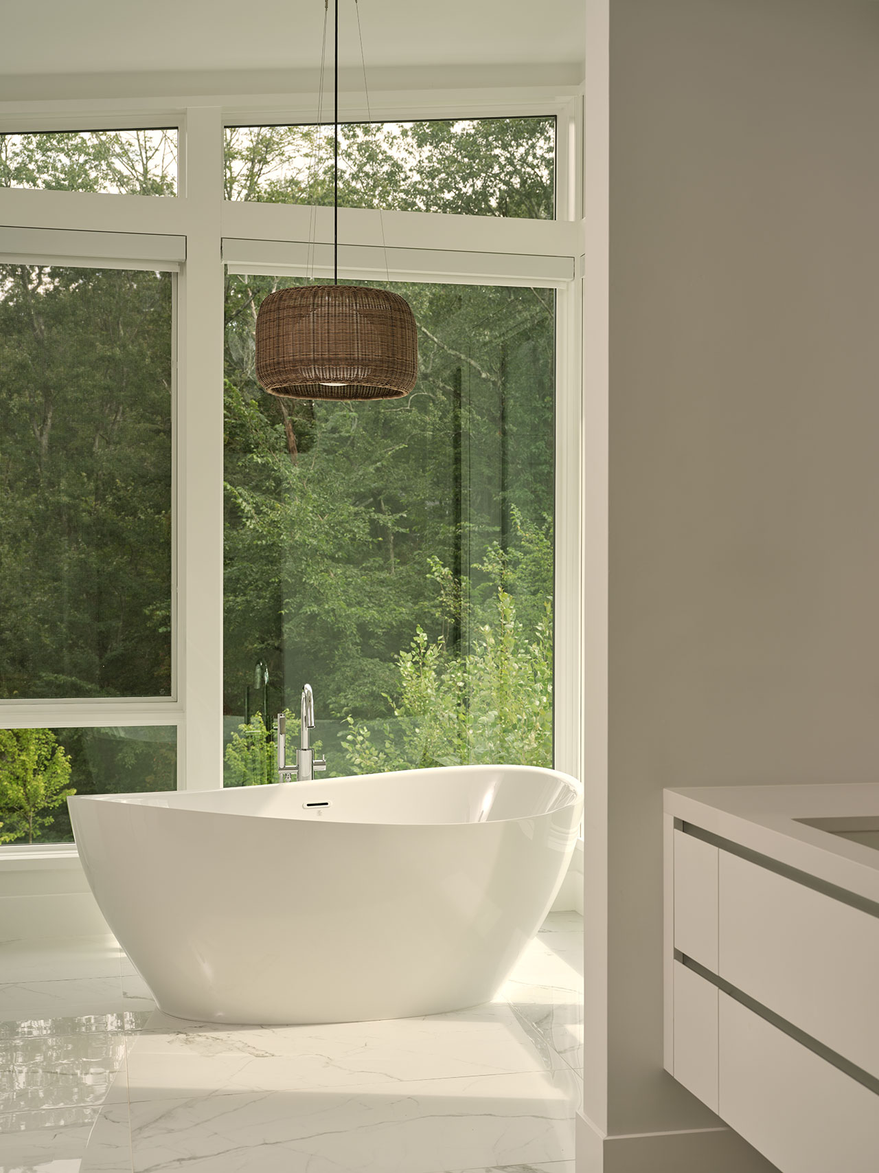 A modern bathroom with a white freestanding bathtub near large windows overlooking greenery, a wicker pendant light above, and a white vanity on the right.