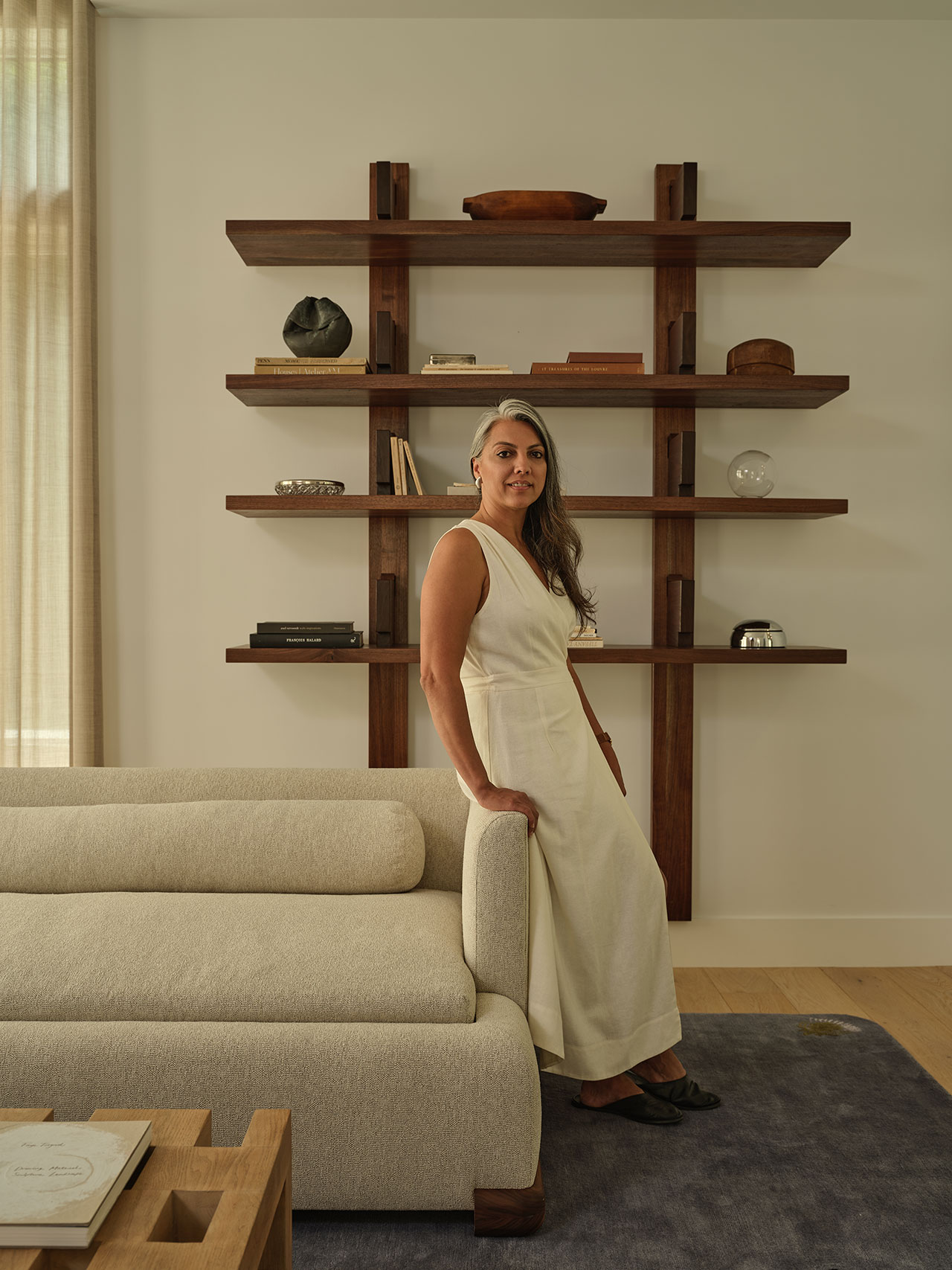 A woman in a white dress stands beside a light-colored sofa in a modern living room with wooden shelves and decorative objects on the wall behind her.