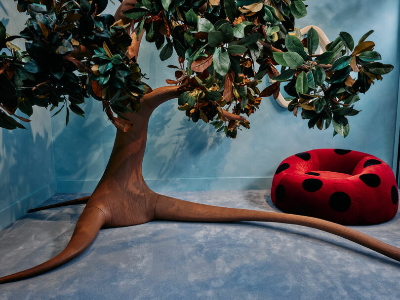 A large, sculptural artificial tree with broad leaves is next to a red, ladybug-patterned chair on a blue carpet in a minimal room.