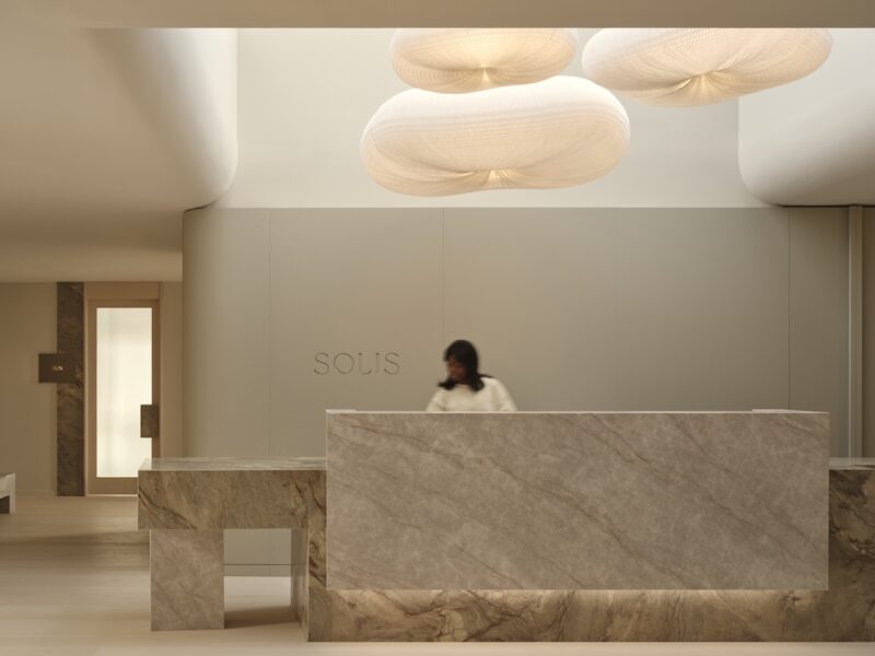 A person stands behind a marble reception desk in a modern, minimalist lobby with soft lighting and sculptural ceiling lights.