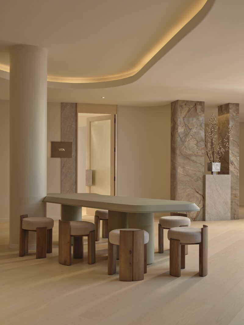 Minimalist interior with a long green table, six wooden stools, curved ceiling lighting, and neutral-toned walls accented by marble columns and a floral arrangement.