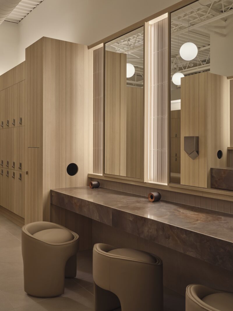 Modern locker room with wooden lockers, marble vanity countertop, round mirrors, and upholstered stool seating under soft ambient lighting.