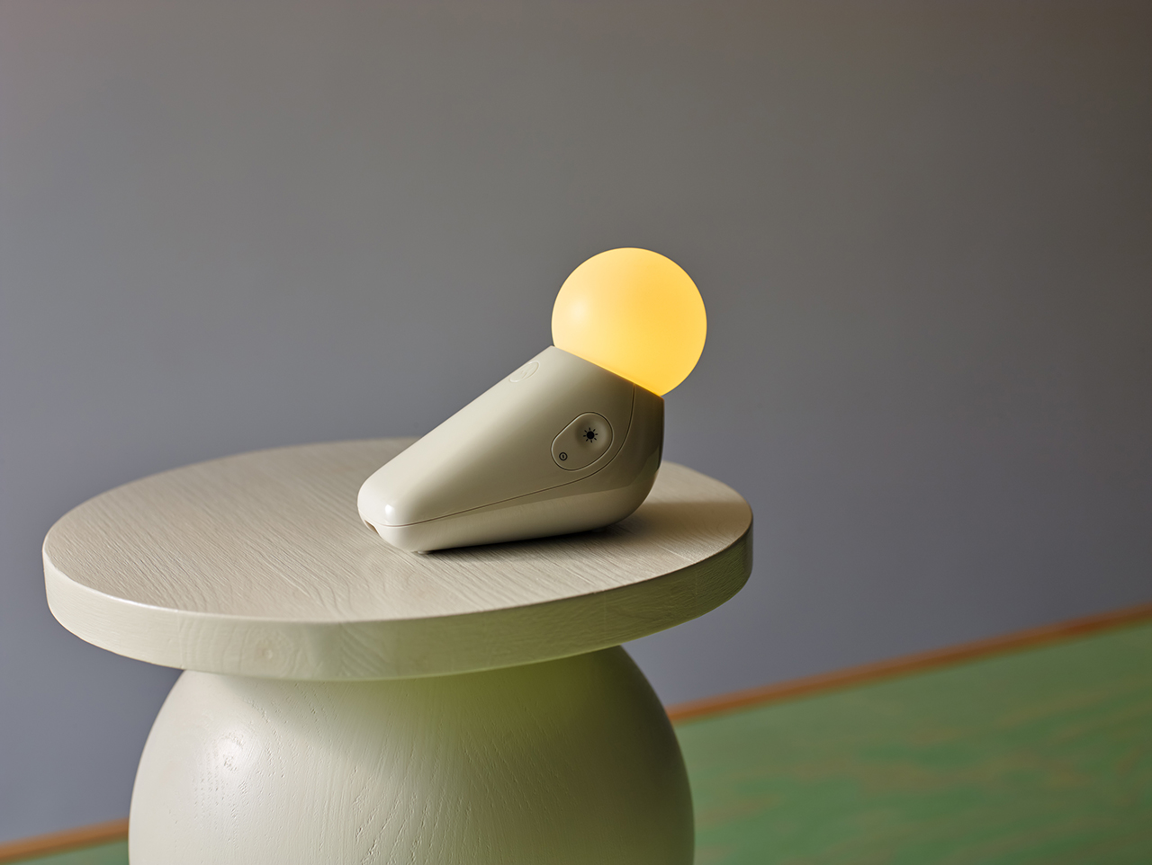 A small, modern lamp with a round yellow light and angled base sits on a round, light-colored wooden table against a plain background.