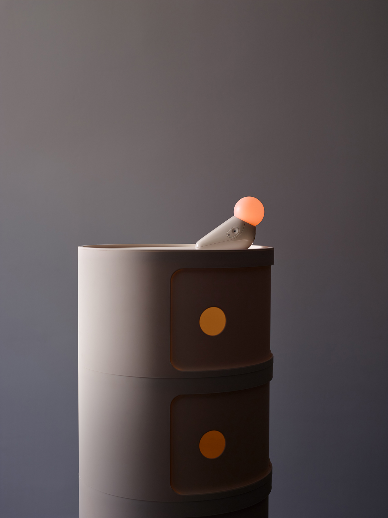 A small, cylindrical object with a glowing orange tip sits atop a two-drawer cabinet with round yellow handles against a plain, dark background.