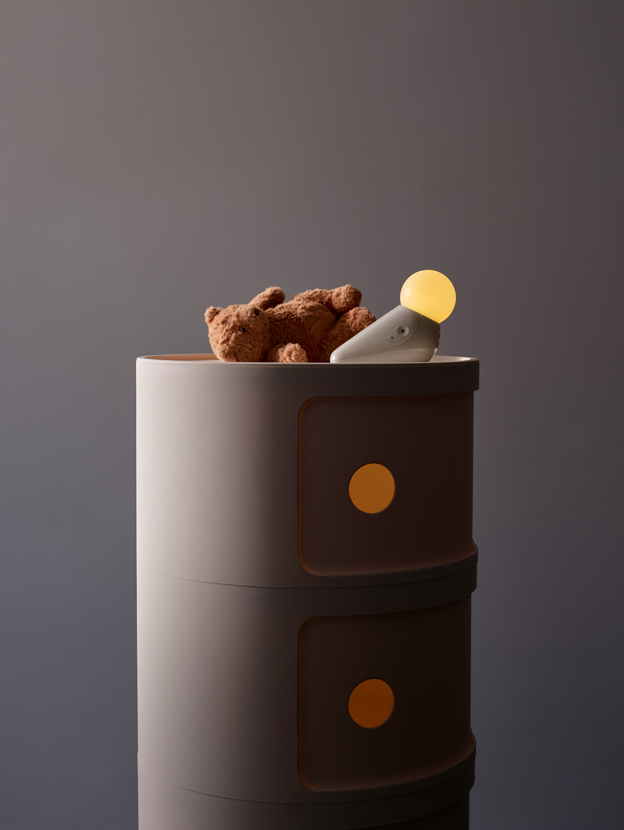 A small brown teddy bear and a modern bedside lamp with a round bulb sit on top of a round, two-drawer cabinet with yellow circular handles.