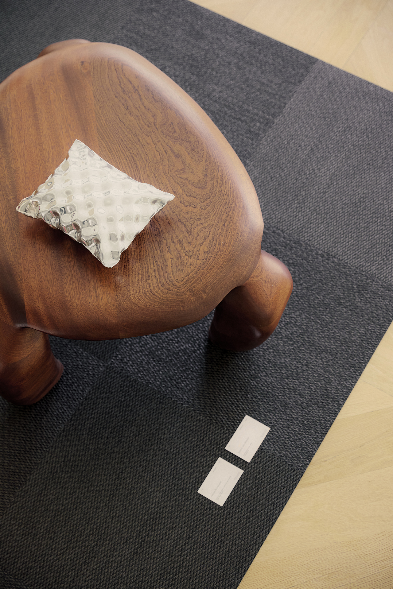 A small, sculpted wooden stool with a textured glass object on top sits on a dark textured rug, with two dgert Comms business cards placed nearby on the floor.