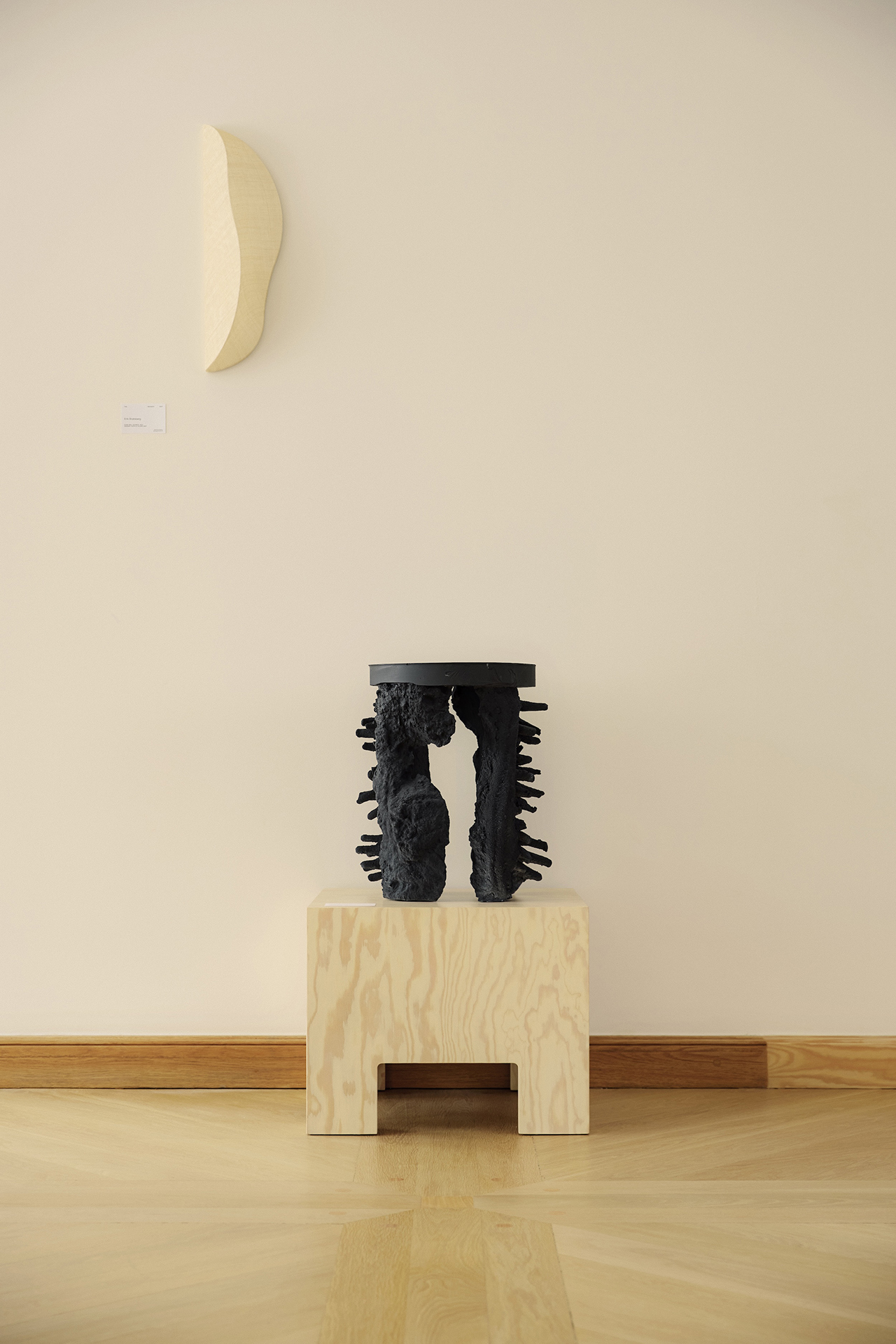 A black abstract sculpture with rough edges sits on a light wooden pedestal, part of the dgert Comms collection, beneath a crescent-shaped wall art on a cream-colored wall.