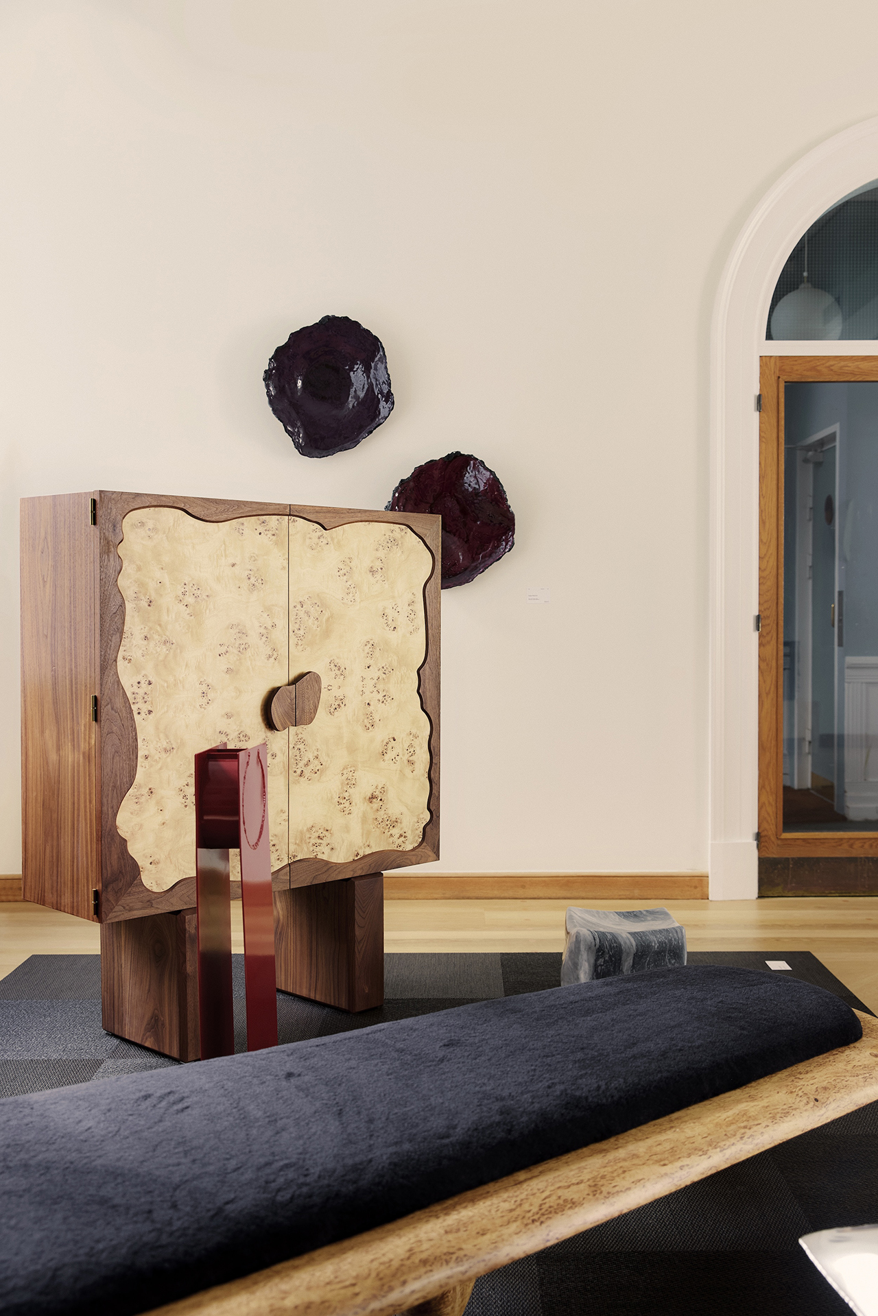 A wooden cabinet with a light-toned, irregular front by dgert Comms stands beside modern decor, including wall art and a low bench, in a minimalist, well-lit room with wood flooring.