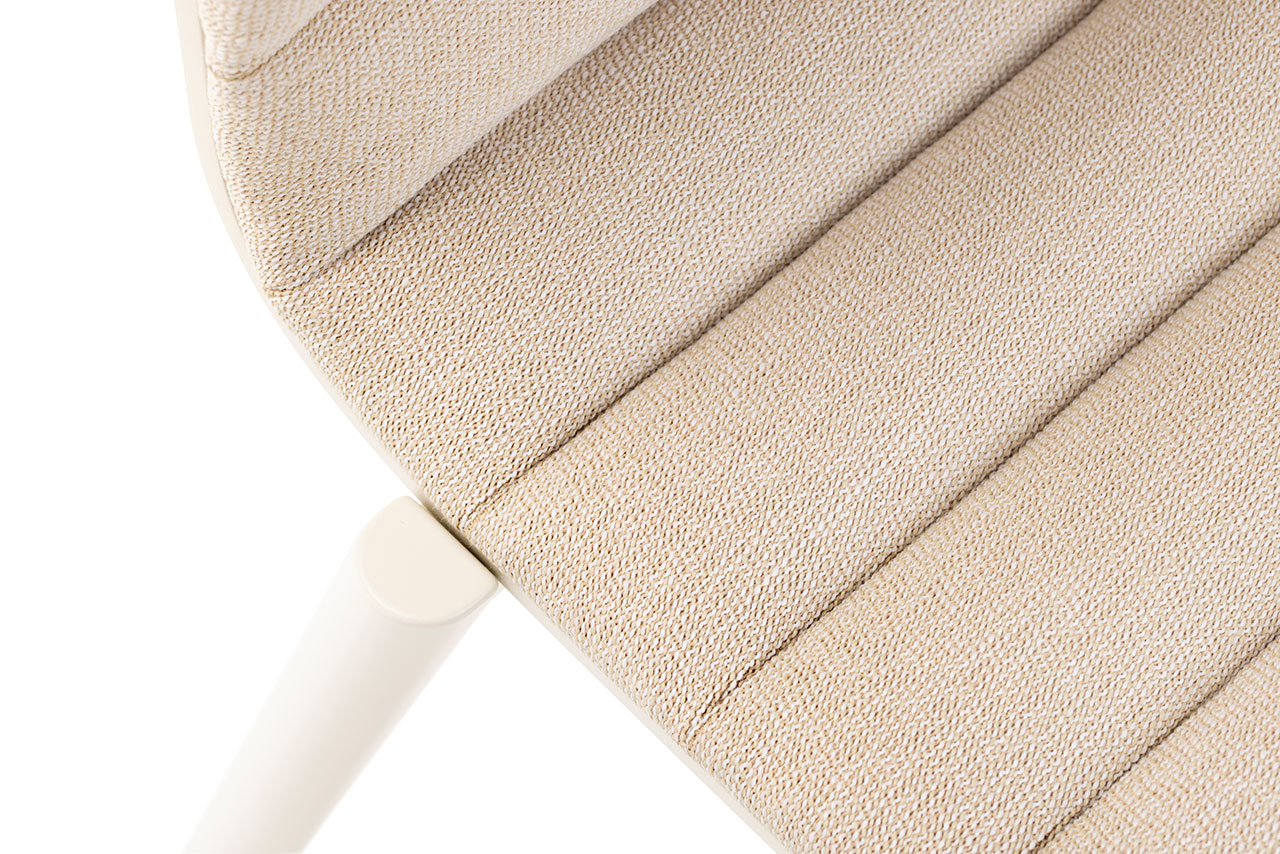 Vista de cerca de una moderna silla tapizada en beige diseñada por Alexander Gufler con tela texturizada y costuras horizontales en el asiento, parte de una pata blanca visible.
