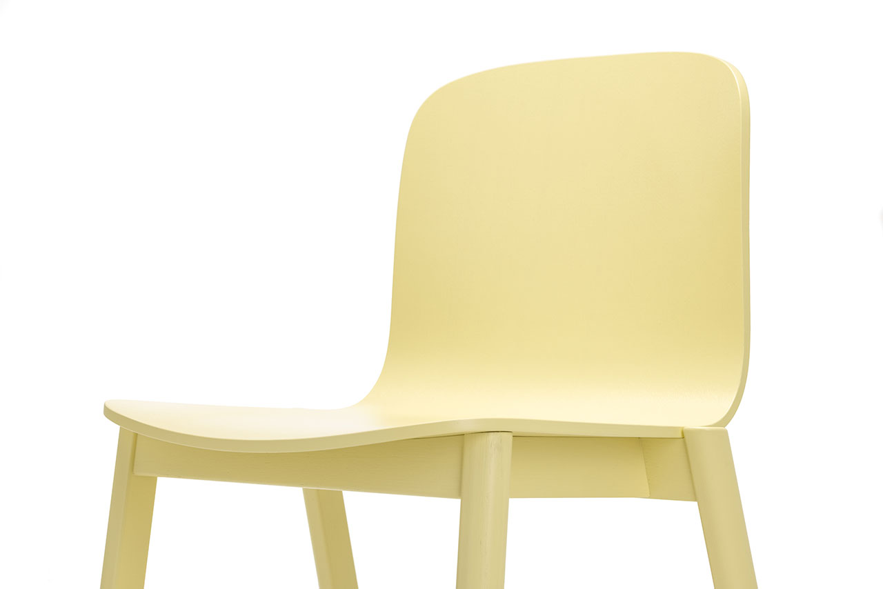 La silla minimalista de madera de color amarillo pálido de Alexander Gufler presenta un respaldo y un asiento curvos sobre un fondo blanco cuando se ve desde un lado.