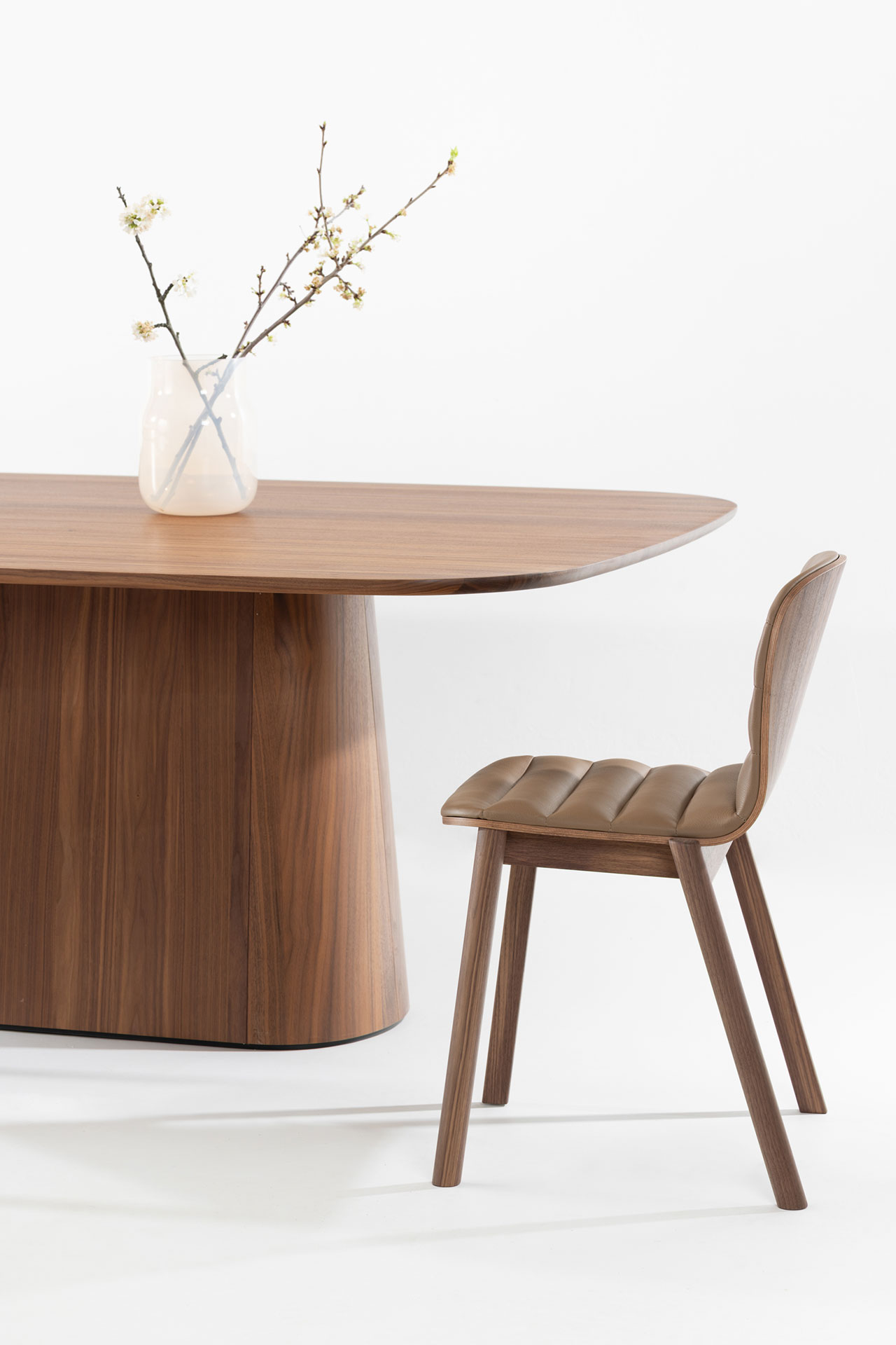 Junto a la mesa del comedor de madera se encuentra una silla de madera con asiento tapizado diseñada por Alexander Gufler. Sobre la mesa hay un jarrón blanco con ramas en flor.