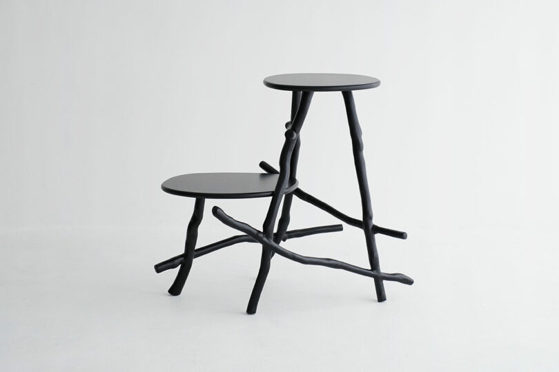 Two black tables with uneven, branch-like legs and circular tops are connected together, set against a plain white background.
