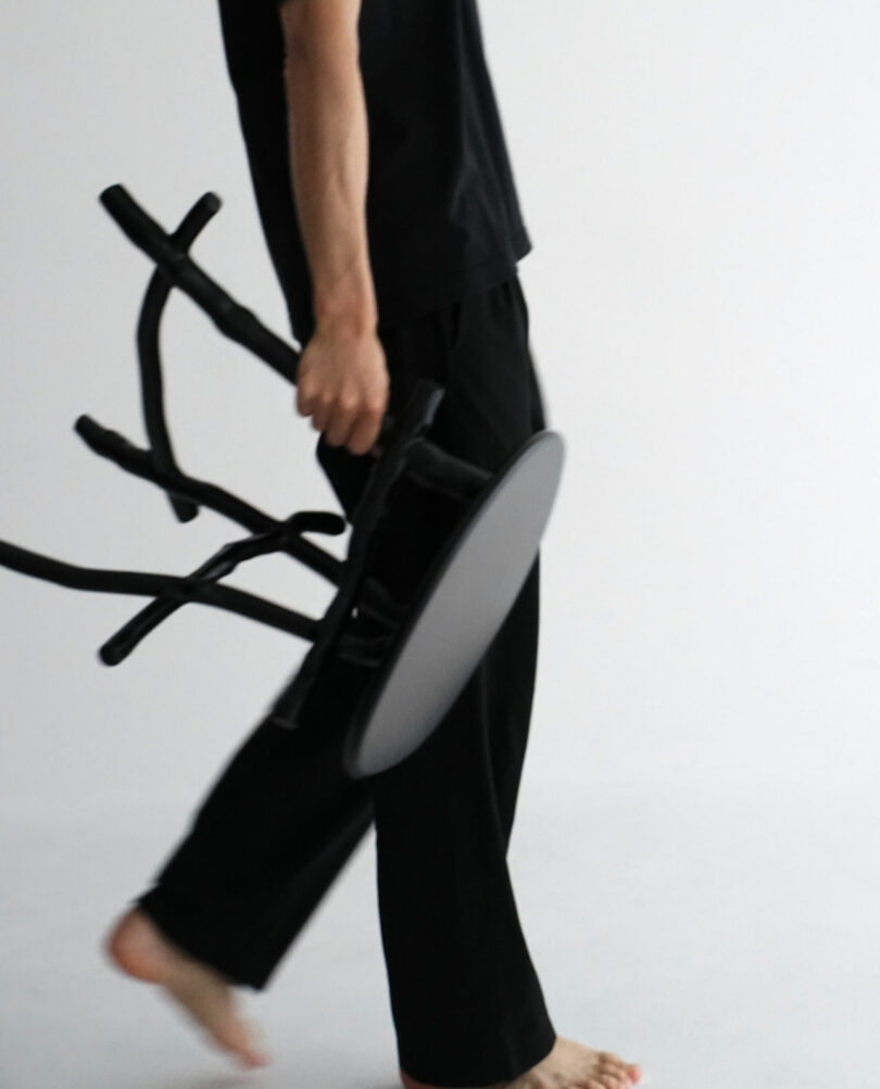 A barefoot person dressed in black pants and a black shirt holds a black stool while walking across a plain, light-colored floor.