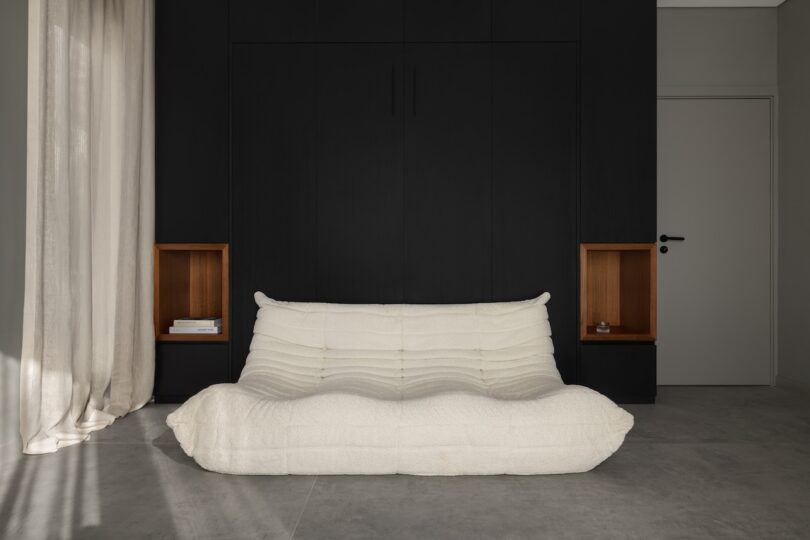 A modern minimalist room with a cream-colored, plush sofa in front of a black built-in cabinet, flanked by wooden shelving alcoves, and light curtains on the left.