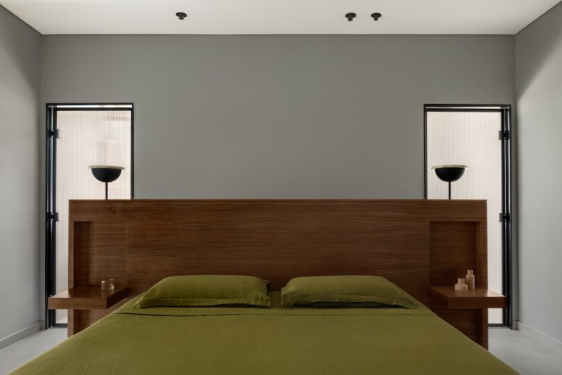 Minimalist bedroom with a wooden headboard, olive green bedding, two small lamps, and two windows on either side of the bed.
