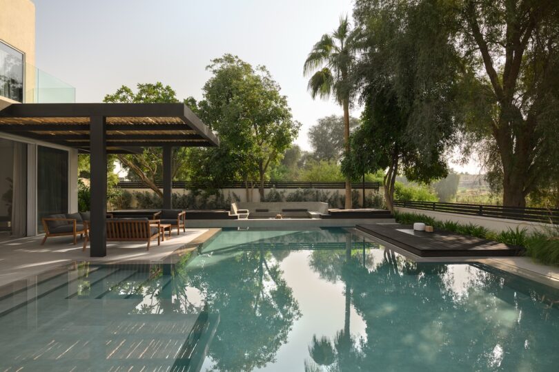 Modern outdoor patio with wooden pergola, seating area, and large swimming pool surrounded by trees and greenery on a sunny day.
