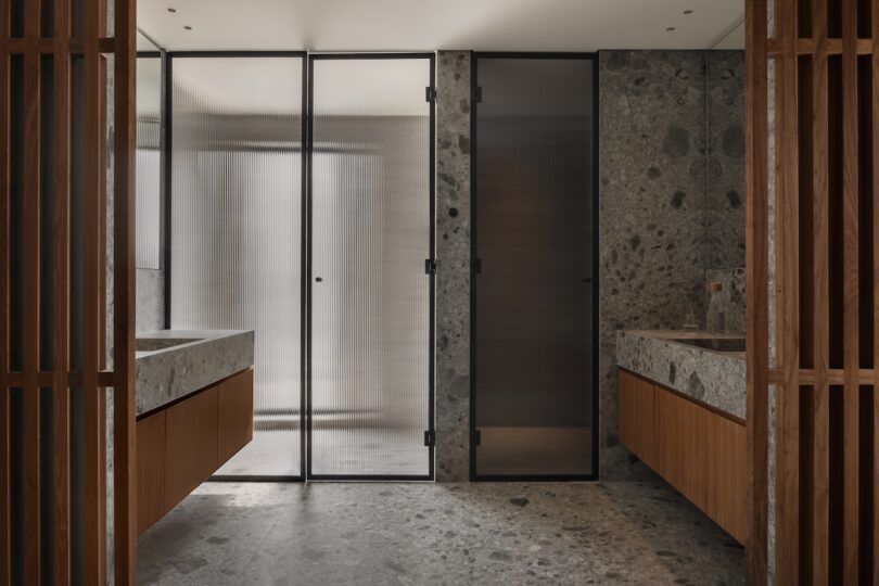 Modern bathroom with stone floors and countertops, wooden cabinetry, and frosted glass doors dividing spaces, viewed from the entrance.