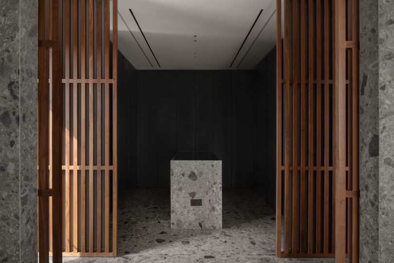 A minimalist interior with stone floors and walls, wooden slatted doors, and a central stone pedestal under a ceiling with recessed lighting.