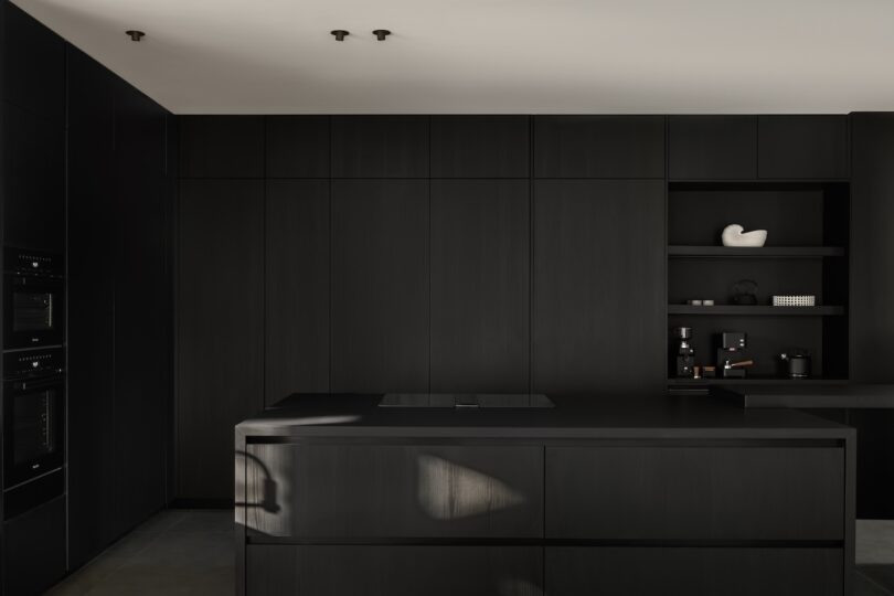 Minimalist kitchen with matte black cabinetry, an island counter, built-in ovens, and open shelves holding decorative objects and kitchenware.
