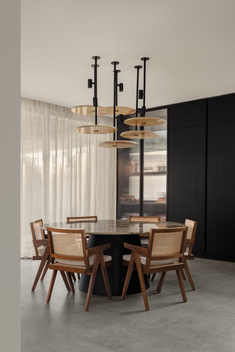 A modern dining room with a round table, six wooden chairs with cane backs, floor-to-ceiling curtains, black wall panels, and a contemporary pendant light fixture.