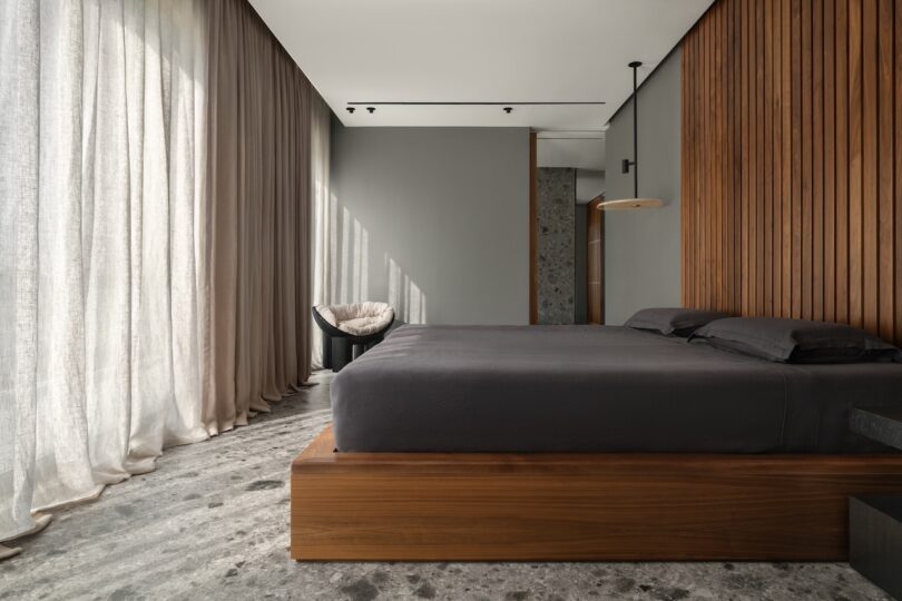Minimalist bedroom with a wooden platform bed, gray bedding, floor-to-ceiling curtains, and a gray chair in the corner. Vertical wood paneling covers one wall.