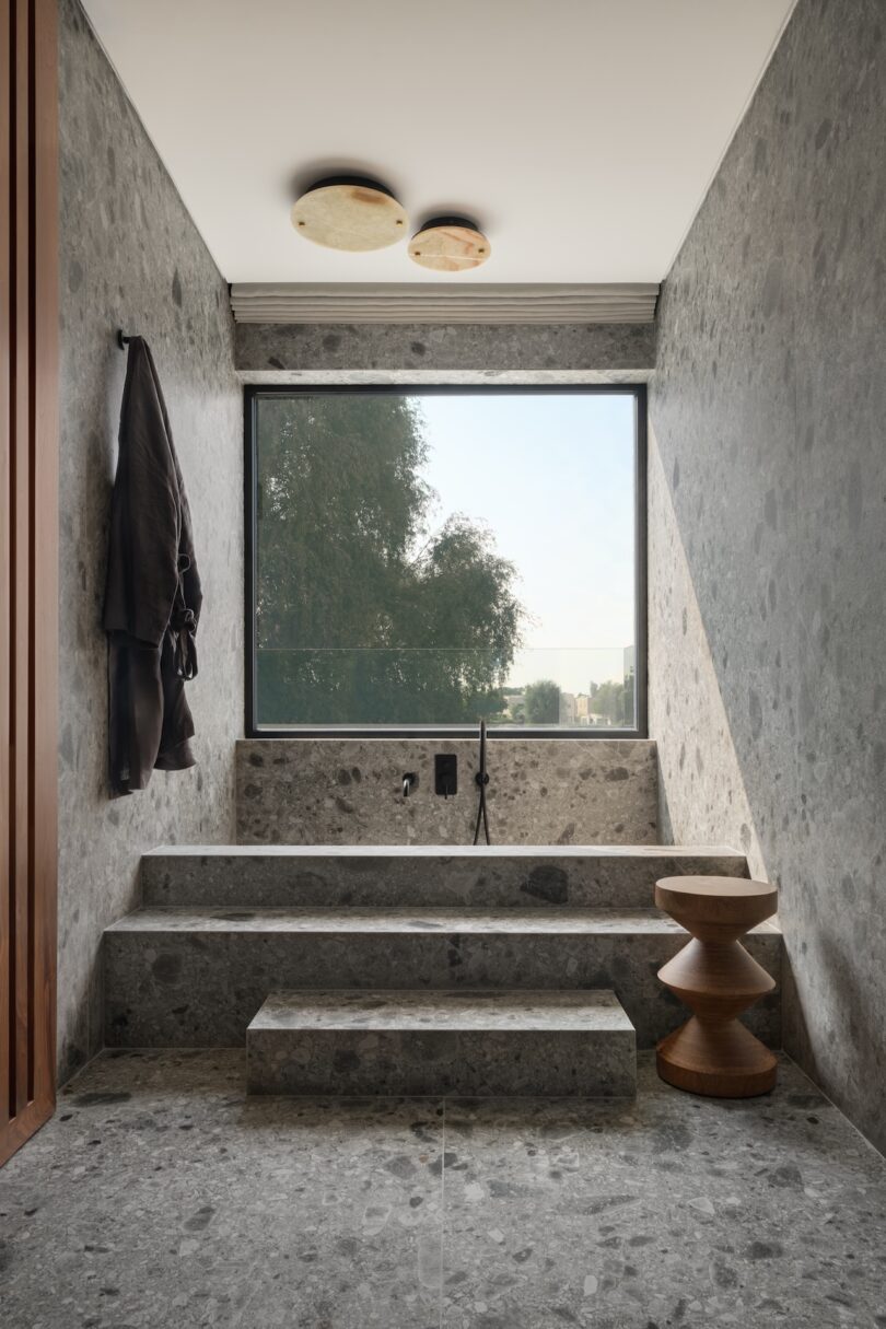 A minimalist stone bathroom features a deep soaking tub with steps, a large window, a robe on the wall, and a wooden stool.