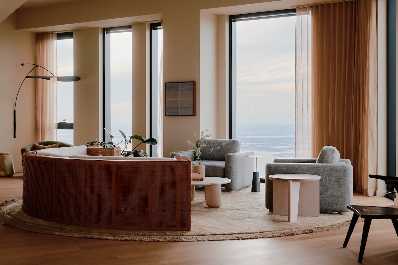 Salón moderno con grandes ventanales, sofá curvo, dos sillones, alfombra redonda, mesa de centro, plantas de interior y decoración en tonos neutros. La luz natural llena todo el espacio.