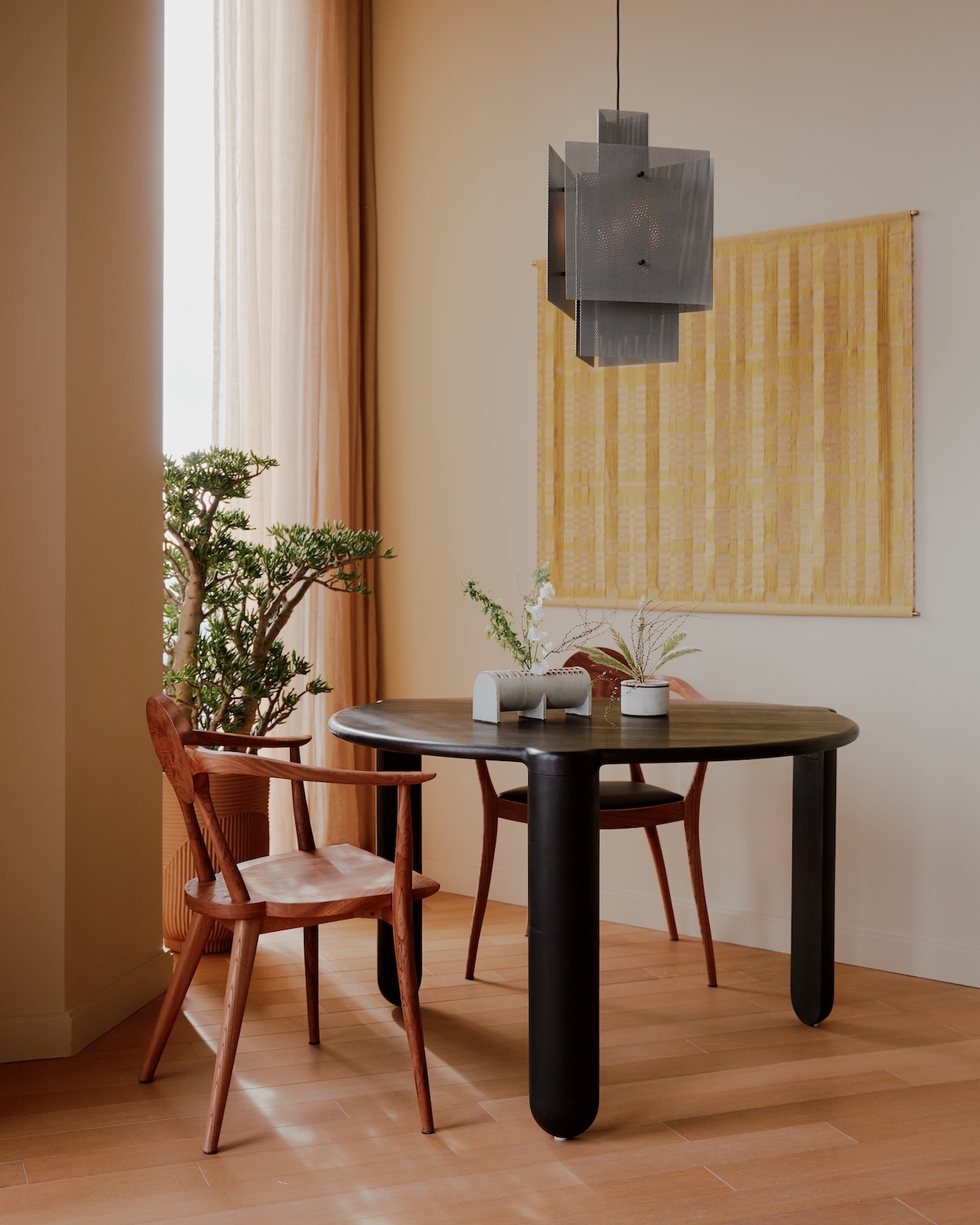 Una mesa redonda negra con dos sillas de madera, dos pequeñas plantas y una decoración minimalista se ubica cerca de un gran ventanal con cortinas transparentes y un tapiz amarillo.