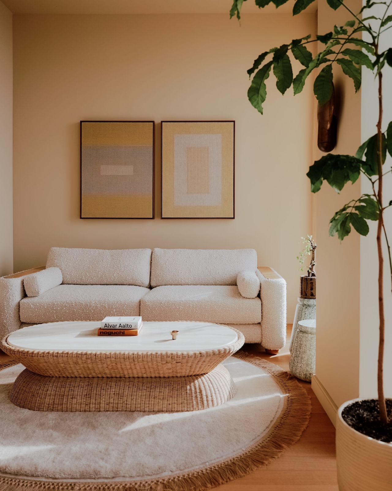 La sala de estar minimalista cuenta con un sofá beige, dos piezas de arte abstracto en las paredes, una mesa de centro redonda tejida y una gran maceta junto a la ventana.
