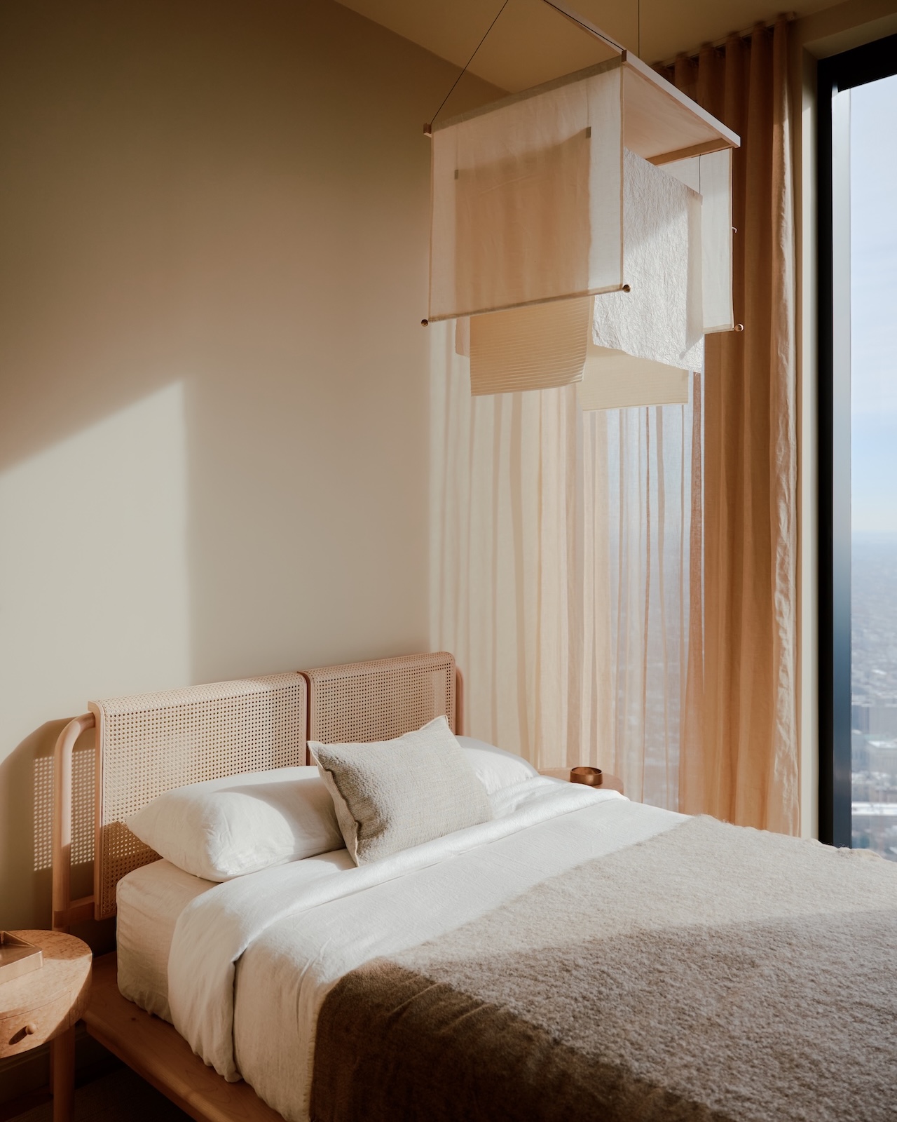 Un dormitorio minimalista con una cama de madera, ropa de cama blanca, manta beige, cortinas transparentes y una moderna lámpara colgante junto a un gran ventanal con vistas a la ciudad.