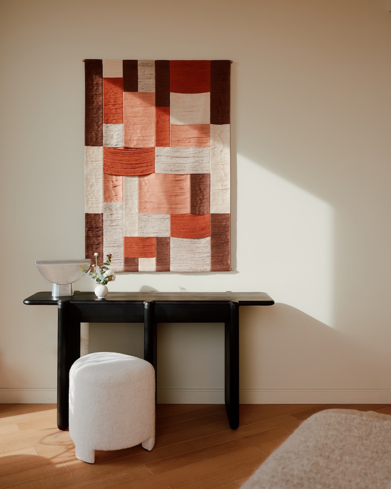 Una pared textil geométrica en tonos rojos, crema y marrón cuelga sobre una mesa consola negra, con un cuenco plateado, un jarrón pequeño y un taburete blanco.