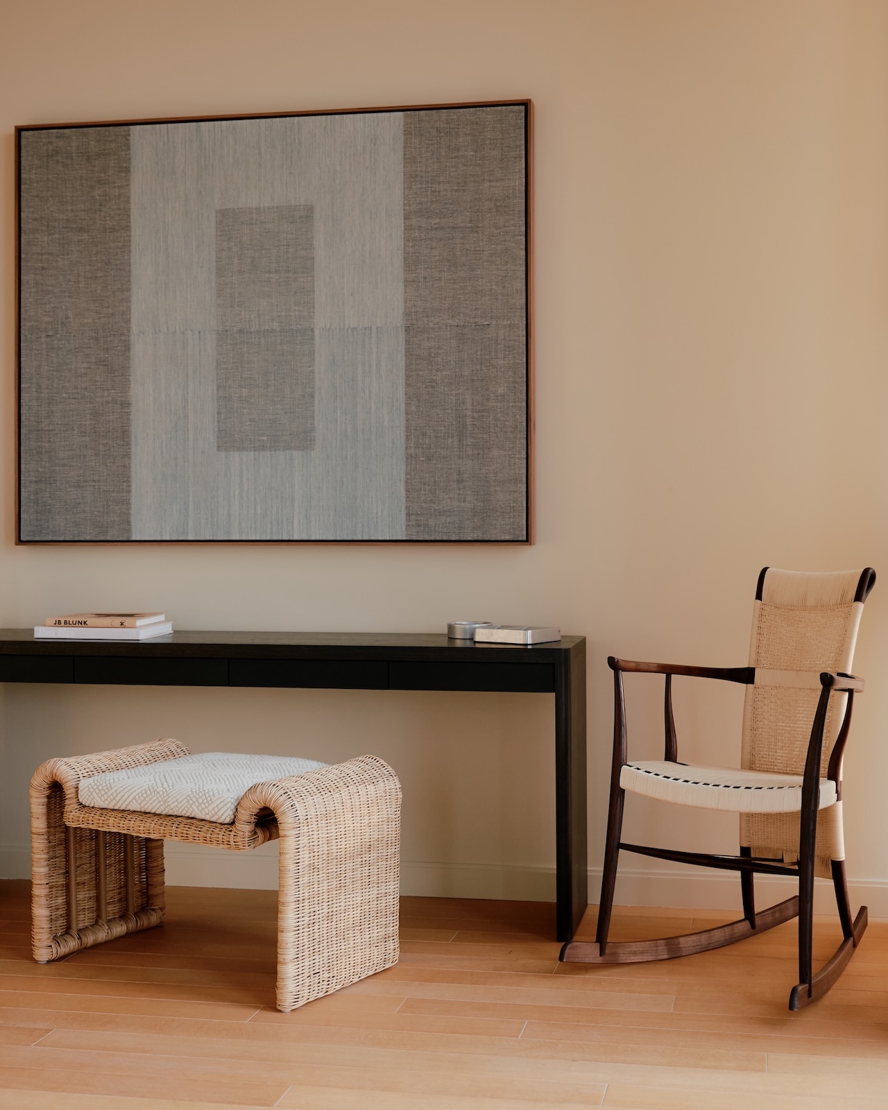 Habitación minimalista con escritorio negro, taburete de mimbre, mecedora beige y gran arte abstracto en una pared clara.