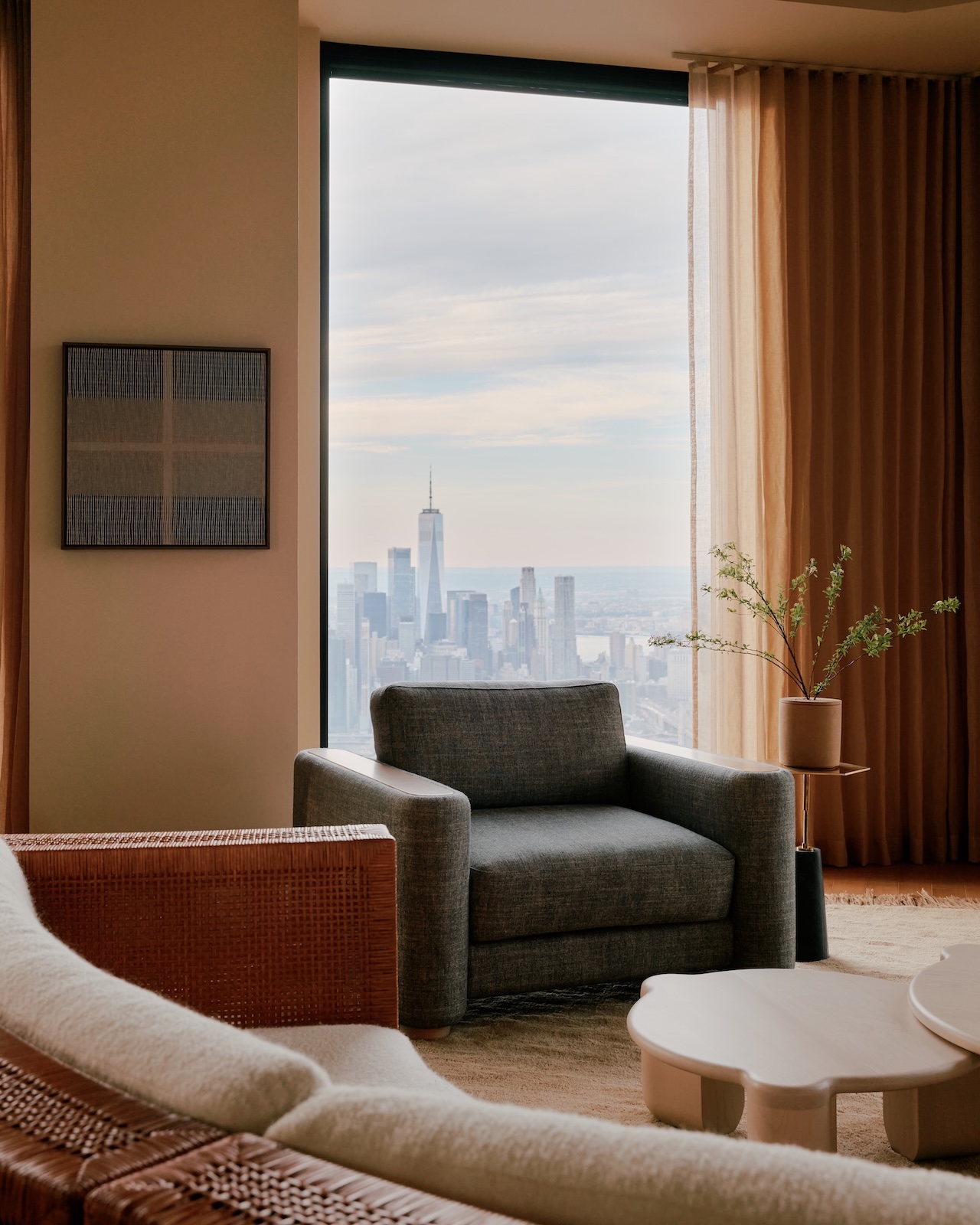Salón moderno con sillones grises y grandes ventanales con vistas al horizonte de la ciudad, incluido un alto rascacielos bajo un cielo nublado.