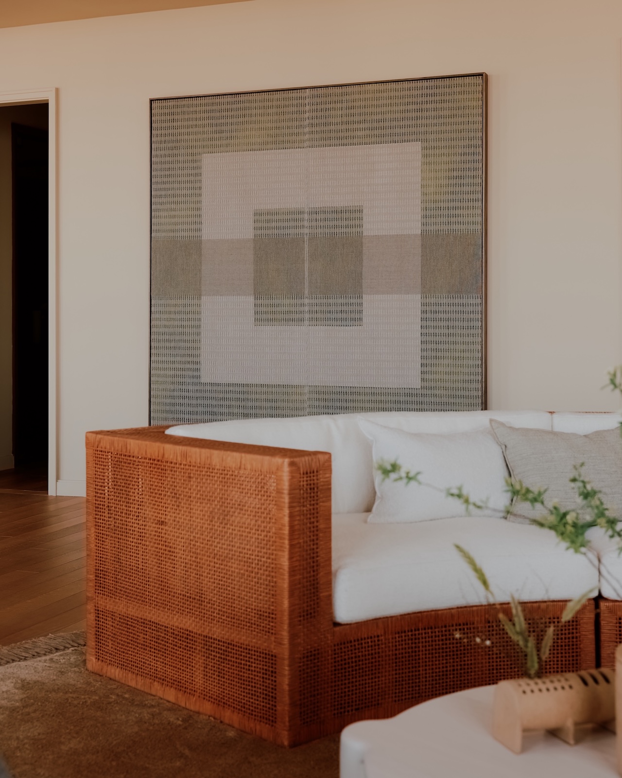 Salón moderno con sofá de ratán, cojines blancos, grandes cuadros geométricos en la pared, suelo de madera y una pequeña mesa con plantas.