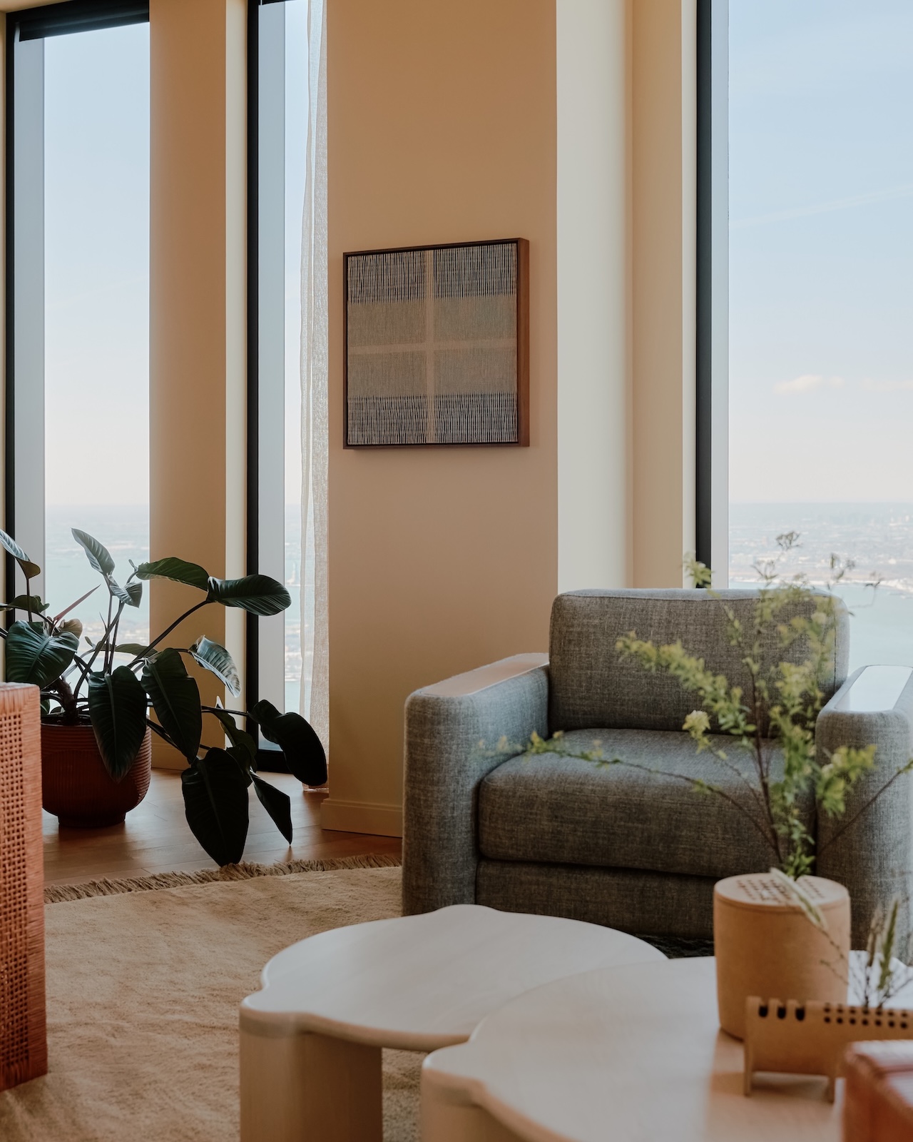 Salón moderno con sillones grises, mesa de madera clara, plantas en macetas y grandes ventanales con vistas a la ciudad y al agua.