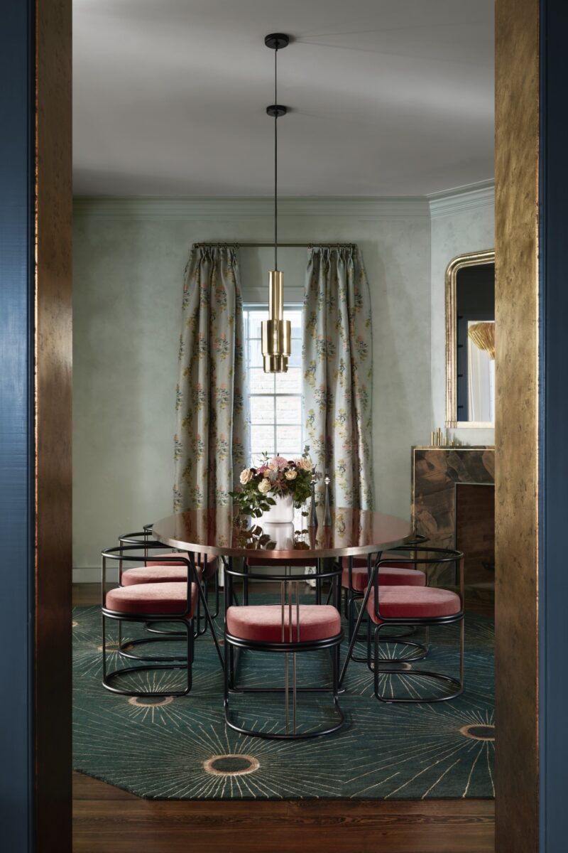 A modern dining room with a round wooden table, six chairs with pink seats, a floral centerpiece, green rug, floral curtains, and a gold pendant light.