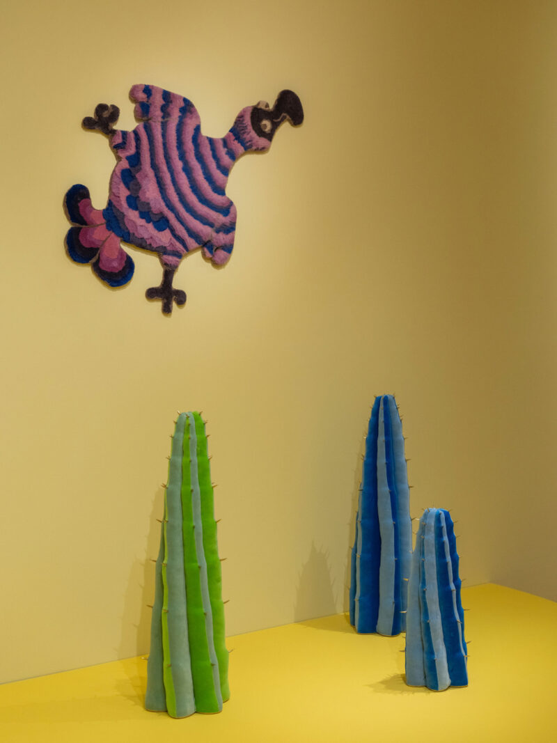 Una colorida obra de arte de un pájaro a rayas cuelga de la pared amarilla, y tres esculturas verticales de cactus estilo Haas Brothers Uncanny Valley en verdes y azules cuelgan del piso amarillo.