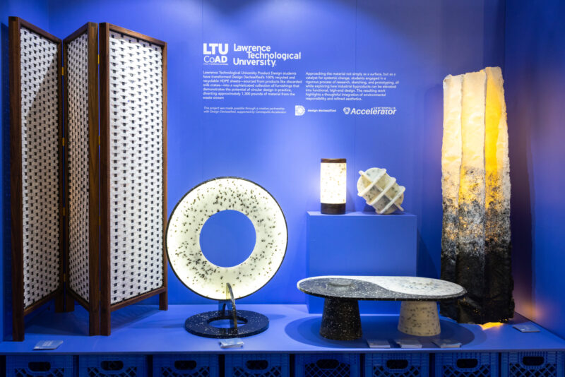 A blue-walled exhibit displays modern furniture and lighting pieces made of light and dark materials, including a lamp, table, screen, and decor items, with Lawrence Technological University signage.