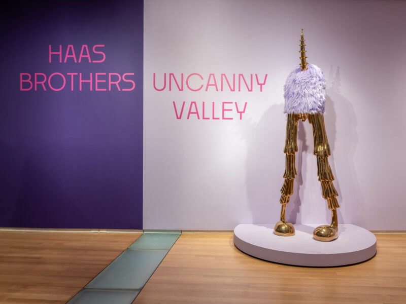 una escultura peluda de cuatro patas dorada y blanca se alza sobre una plataforma circular junto a la pared de lectura "Valle inquietante de los hermanos Haas" Las letras rosas reflejan el estilo caprichoso de la serie Uncanny Valley de Haas Brothers.