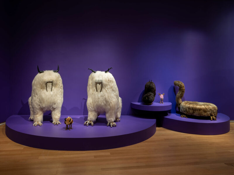 Cinco esculturas de animales de fantasía a gran escala, con rasgos y pelaje exagerados que son característicos de la estética del valle inquietante de los hermanos Haas, se exhiben sobre plataformas moradas contra paredes moradas en el fondo de la galería.