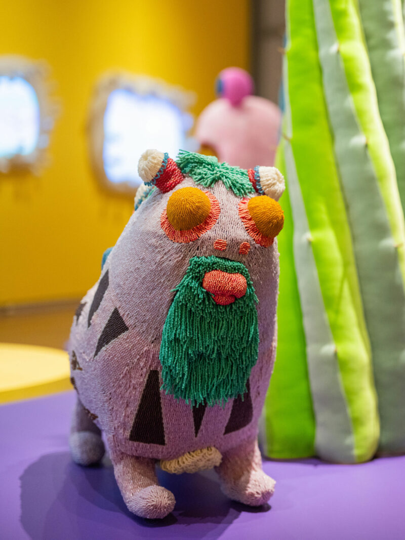 Contra la superficie violeta se encuentra una colorida escultura cubierta de hilo de un animal con barba verde y ojos anaranjados, junto a una estructura verde parecida a un cactus que evoca el estilo caprichoso del Uncanny Valley de los hermanos Haas.