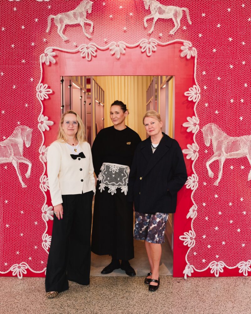 Three women stand in front of a red wall decorated with white lace horses and a framed doorway, posing for a group photo indoors, each showcasing chic Baum und Pferdgarten-inspired style.