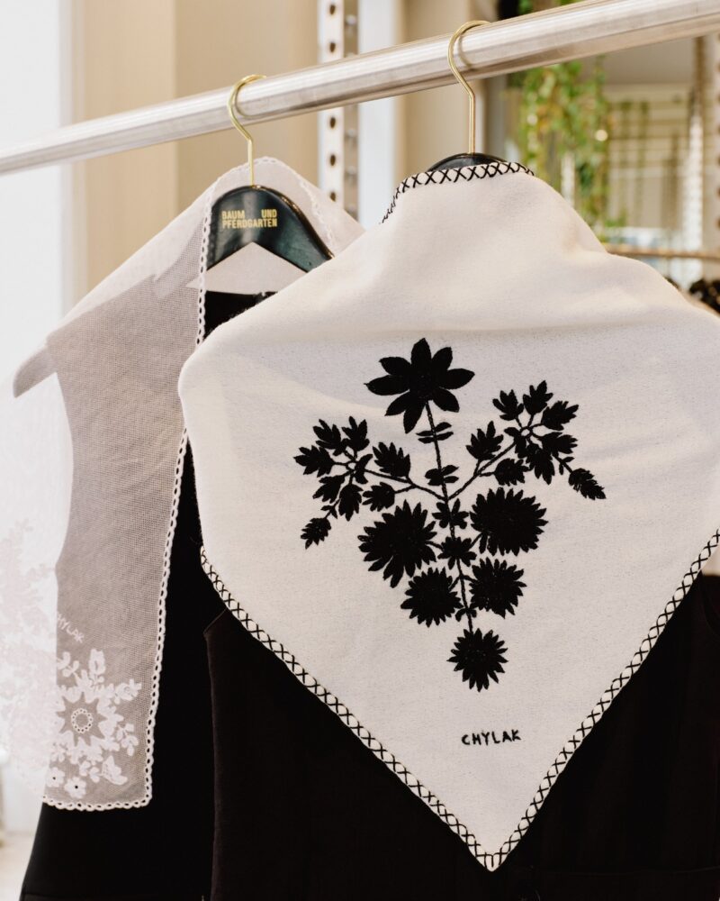 Two black garments on hangers, each with a white cloth draped over the shoulders; one cloth features black floral embroidery and the name "Chylak".