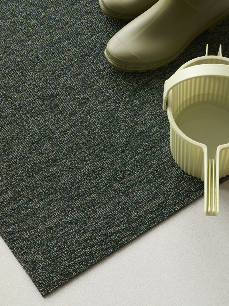 Botas de goma verdes y cubos de plástico de color verde claro se asientan sobre una alfombra texturizada de color verde oscuro que se asienta sobre un piso claro, una disposición que evoca el estilo clásico de Martha Stewart con un toque de practicidad de Hachiman.