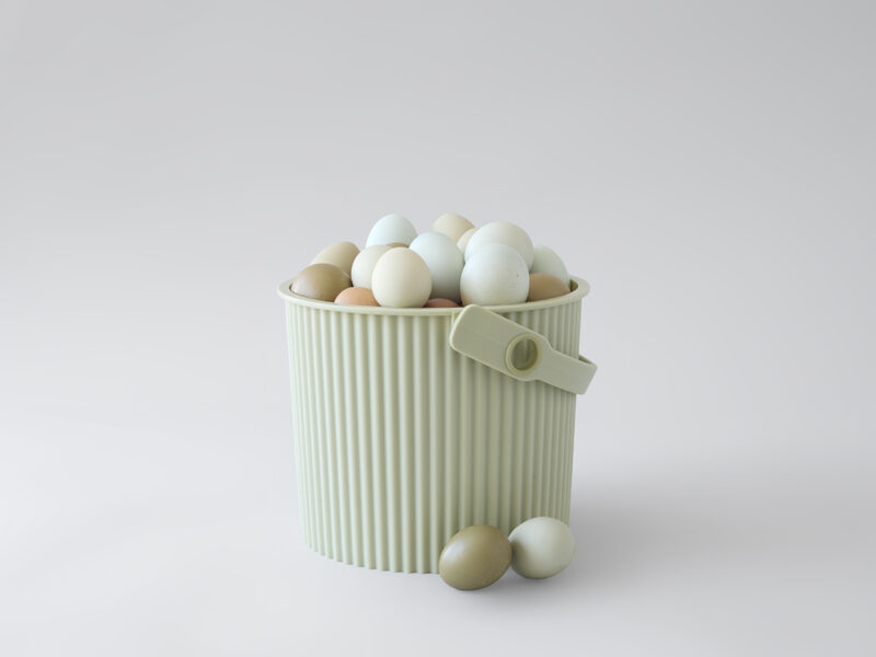 Un cubo acanalado de color verde claro (que recuerda el estilo característico de Martha Stewart) se encuentra sobre una superficie blanca llena de huevos de colores, con algunos huevos y una pinza para la ropa cerca.