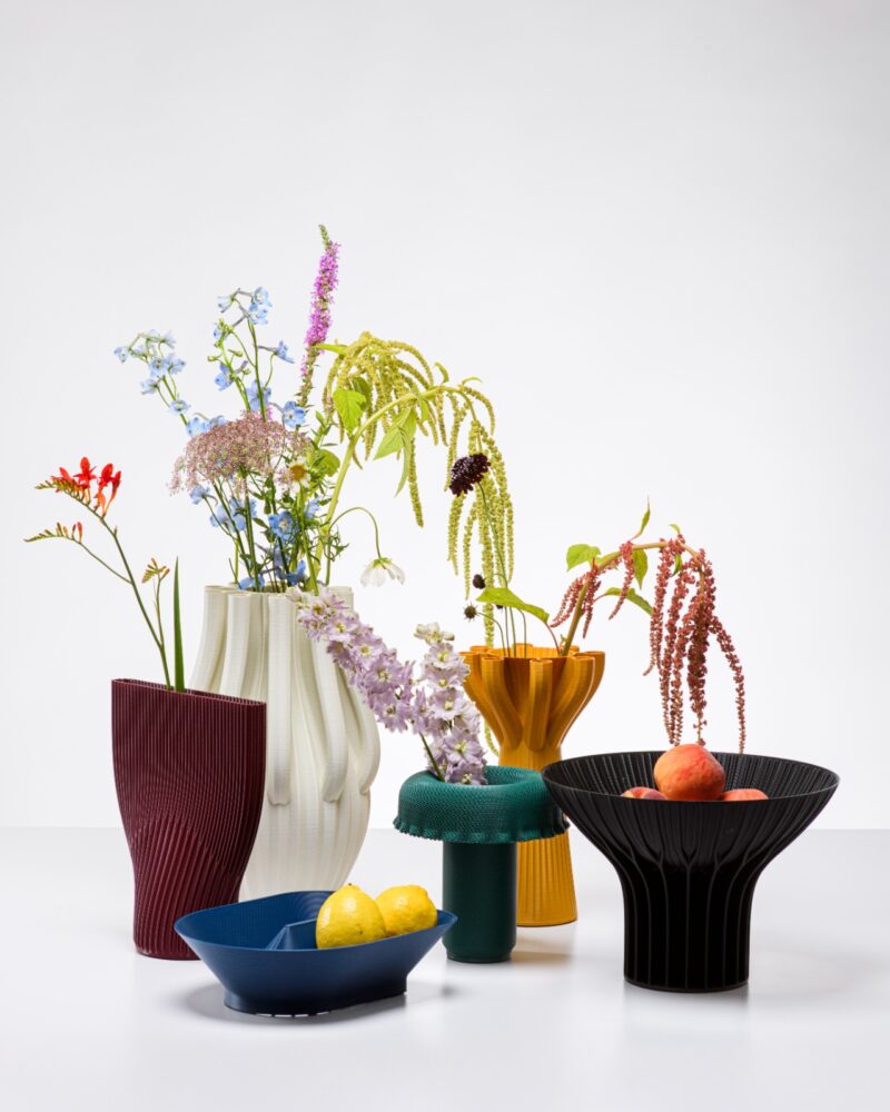 Six colorful vases hold assorted flowers, alongside a blue bowl with lemons and a black bowl with peaches, all arranged on a white surface with a plain white background—an inviting display that perfectly complements sustainable home furnishings.