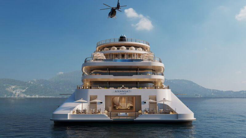 Rear view of a large luxury yacht named "Amangiri" docked on calm water, with a helicopter flying overhead and green hills in the background under a clear sky.